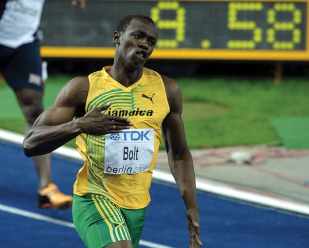 Photo of Usain Bolt patting his chest in a gesture of humility while walking along running track with "9.58" visible on results board behind him