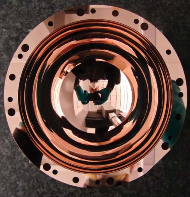 Photo of a concave copper hemisphere, with bolt holes around its rim, which is so shiny it reflects the camera and photographer