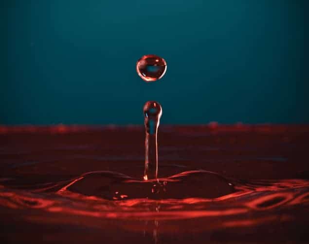 Photograph of a droplet of liquid mid-action in the air, with ripples in the liquid bath beneath it.