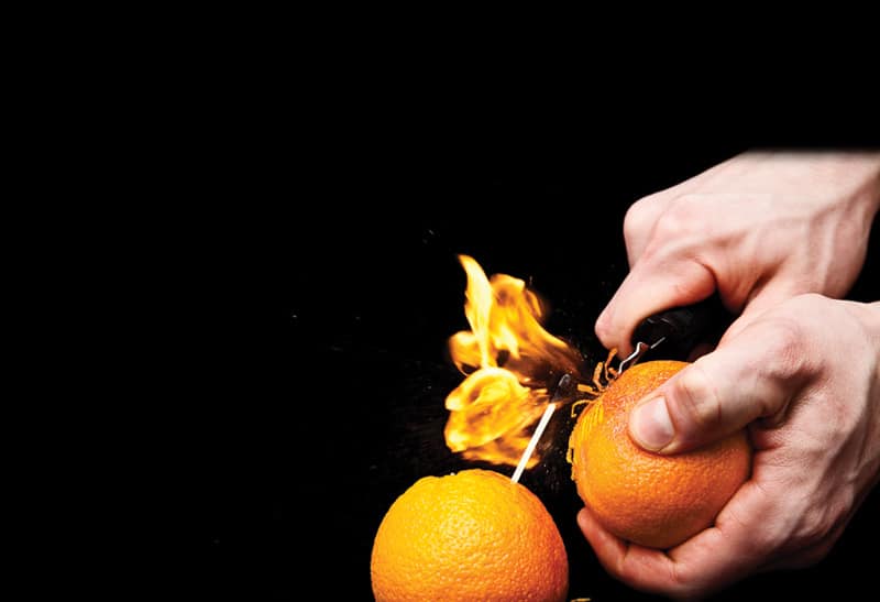 A man's hands holding an orange and setting oil in the orange peel on fire