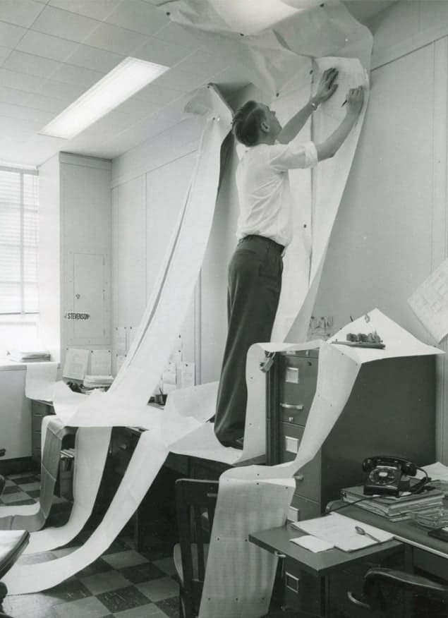 Black-and-white photo of a scientist, Russell White, standing on a desk trying to read a very long computer printout at the RAND think tank in the 1950s