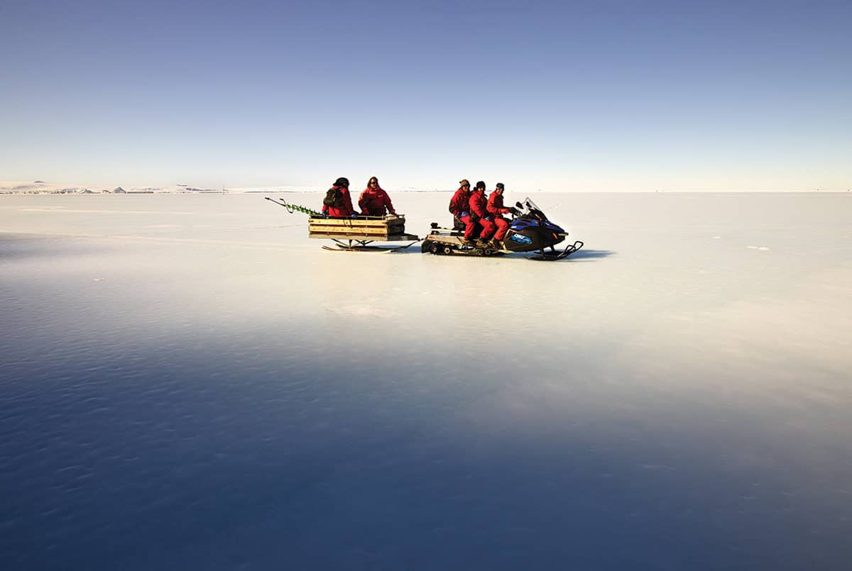 Wide snowy landscape with two snowmobiles ridden by figures dressed in red