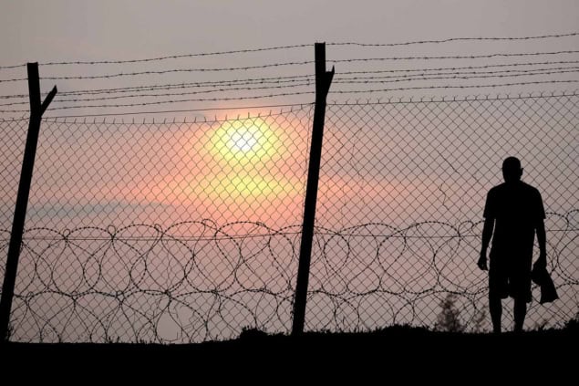 pwaug14-moskvitch-fence-hero Man standing behind a barbed wire fence at sunset