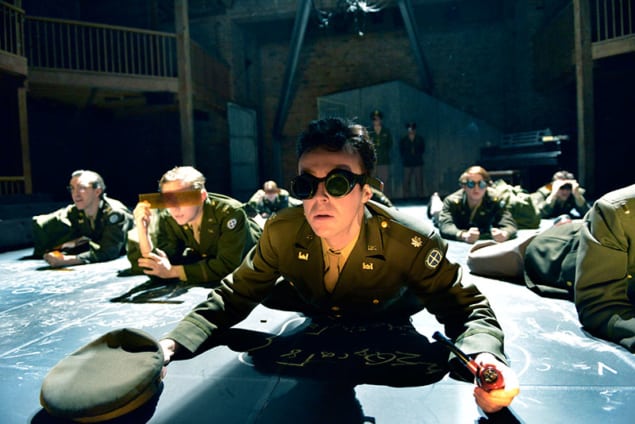 Cast members in the play Oppenheimer lie prone on stage wearing dark glasses as they await the first atomic bomb explosion