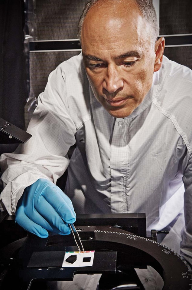 Photograph of a man in a white lab coat and blue gloves holding some tweezers above two dark chunks of material about the size of sugar cubes