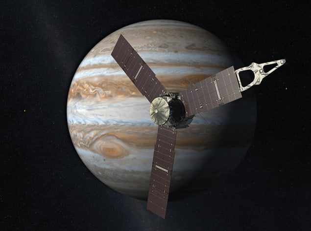 Artistic impression of the Juno spacecraft, consisting of a central gold-hued structure from which emanate three symmetrical long rectangular solar-celled brown arms, positioned in front of Jupiter