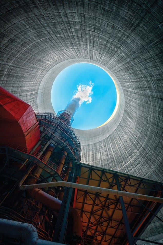 Photo taken from the bottom of a cooling tower, with red and orange machinery in the foreground, steep grey walls, and a circle of clear blue sky, into which a small puff of cloud is escaping