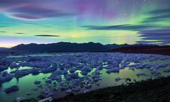 Photo of Iceland's Jokulsarlon glacier lagoon on a night with aurora present. The lagoon is dotted with icebergs that appear purple under the purple-green light of the aurora