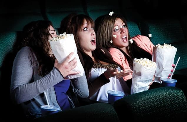 Three people in cinema seats, with popcorn and drinks, have a surprised or scared look on their faces