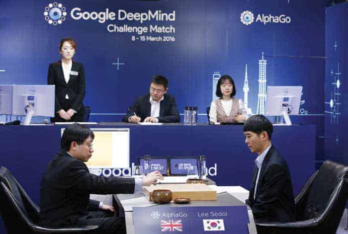 Two men sit opposite each other playing Go on the wooden board between them. The left player, who has a computer screen by him, is marked with a sign as AlphaGo and a Union Jack schuld@ukzn.ac.za the flag of the UK. The man on the left is marked as Lee Sedol, with the flag of South Korea