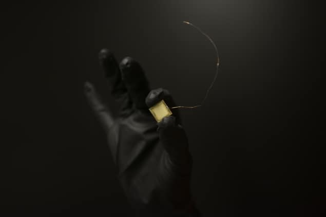 Photo of a gloved hand holding a square chip with a long tail between thumb and forefinger