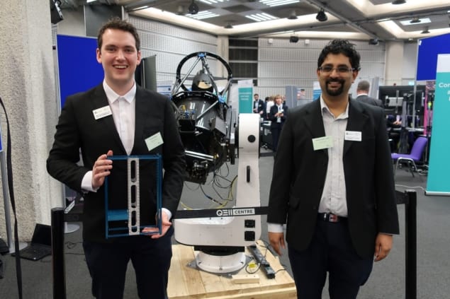 Two men standing and smiling in front of a receiver and holding a metal frame containing a prototype single photon source