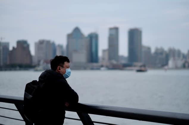 Coronavirus-2019-1211149704-iStock_RobertWei A man wearing a mask stands in front of a body of water with a city skyline in the distance.