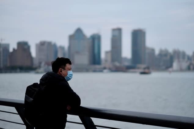 A man wearing a mask stands in front of a body of water with a city skyline in the distance.