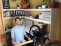 Resty Collado in front of the camera and his laptop, surrounded by shelves full of books, photos and a guitar