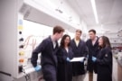 Four students standing in front of a fume cupboard in a laboratory, looking at a lab notebook, as Andrea Armani speaks with them