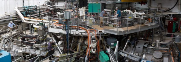 A photograph of the DIII-D tokamak taken before the pandemic. It shows four people in hard hats (but not masks) working closely together on the tokamak.