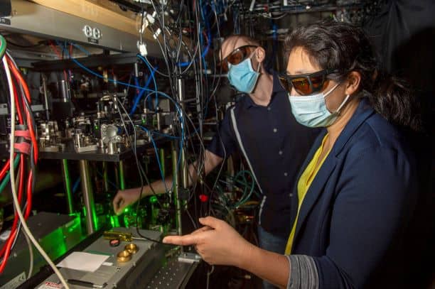 Two researchers in masks stand next to an optical bench containing a laser, mirrors and other equipment.