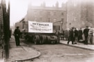 Sepia photograph of a large vehicle carryubg a load wider than the street. To one side and behind are red brick buildings, to the left is a metal fence. Men in suits and in working class clothing stand around the vehicle