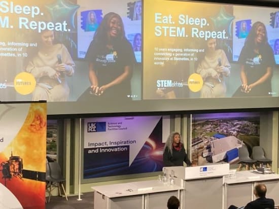 Anne-Marie Imafidon speaking at the Appleton Space Conference, beneath a screen showing photos of smiling young women and the words "Eat. Sleep. STEM. Repeat"