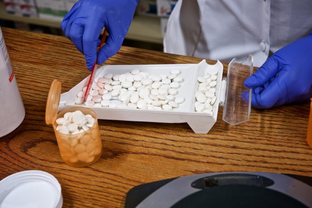 The counting of medicine pills in a pill counter by a pharmacist wearing blue gloves holding a red plastic and pill containers on a wooden counter in a pharmacy
