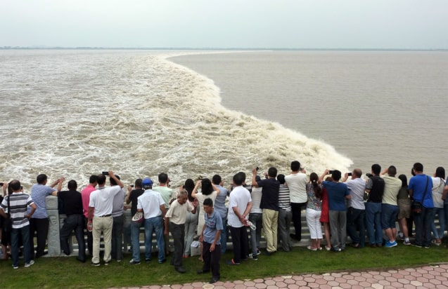 A crowd on the bank of a wide river look at a tidal bore passing