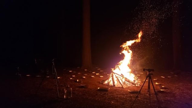 Photo of a small bonfire at night in a forest clearing with flames rising skywards. A camera on a tripod and other equipment are silhouetted against the flames, and aluminium baking trays can be seen scattered around the forest floor near the fire.