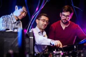 Photo of Nuh Gedik and colleagues Tianchuang Luo and Alexander von Hoegen, pictured in their lab. Gedik is pointing at a piece of laboratory equipment while the other two look on. An electrical cable in the foreground snakes down into the camera's view.