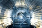 Photo of the Main Spectrometer at the KATRIN experiment during installation of its inner electrode system. The photo is taken inside the experiment and shows a tunnel-like interior with electronics panels covering every visible surface. At the centre is a metal platform with scaffolding and ladders. Three people in clean-room protective clothing are standing on the platform.