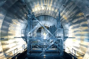 Photo of the Main Spectrometer at the KATRIN experiment during installation of its inner electrode system. The photo is taken inside the experiment and shows a tunnel-like interior with electronics panels covering every visible surface. At the centre is a metal platform with scaffolding and ladders. Three people in clean-room protective clothing are standing on the platform.