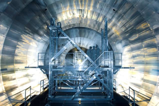 the Main Spectrometer at the KATRIN experiment Photo of the Main Spectrometer at the KATRIN experiment during installation of its inner electrode system. The photo is taken inside the experiment and shows a tunnel-like interior with electronics panels covering every visible surface. At the centre is a metal platform with scaffolding and ladders. Three people in clean-room protective clothing are standing on the platform.