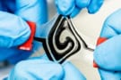 Photo showing four blue-gloved hands pulling at the corners of the battery, which resembles a coil of black gel on a clear plastic surface. The gel and the surface are visibly deformed by being pulled.