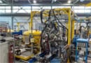 Photo of the set-up in the ISAC-II experimental hall at TRIUMF. The room resembles a high-ceilinged warehouse with metal supports and high windows in the background. In the foreground is an experimental apparatus supported by a yellow metal frame.