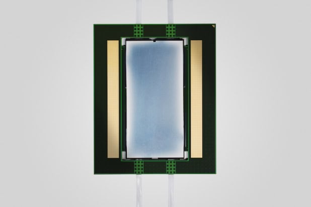 Photo of the Lightmatter device. A long bluish-grey rectangle flanked by thinner gold rectangles is mounted on a larger black rectangle. Wires lead in and out of the device.