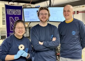 Ezra Marker, Raiden Speelman, and Franz Geiger in front of their spectrometer. They're all smiling and wearing matching navy blue long-sleeve T-shirts with the slogan "this is my laser shirt" written on the chest pocket in light blue. A Northwestern University banner hangs in the background and two monitors displaying graphs and a dashboard can also be seen.
