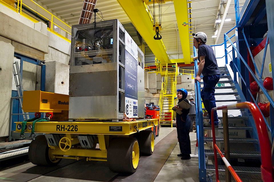 Photo of the BASE-STEP system sitting on a bright yellow trolley after being unloaded from the transport crane, which is visible above it. A woman in a hard hat and head scarf watches from the ground, while a man in a hard hat stands above her on a set of steps, also watching.