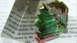 Photo of a jagged, multicoloured, shiny bismuth crystal sitting on a scientific paper