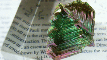 Photo of a jagged, multicoloured, shiny bismuth crystal sitting on a scientific paper