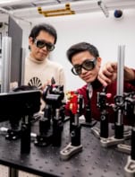 Photo of Jian-Xiang Qiu and Suyang Xua posing with the apparatus they used to detect axion quasiparticles. The apparatus comprises lasers and other components mounted on an optical table. They're wearing laser safety glasses.