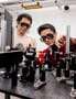 Photo of Jian-Xiang Qiu and Suyang Xua posing with the apparatus they used to detect axion quasiparticles. The apparatus comprises lasers and other components mounted on an optical table. They're wearing laser safety glasses.