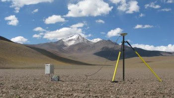 A tripod-shaped structure in a beige-coloured landscape with a snow-streaked, pointy mountain in the bacground
