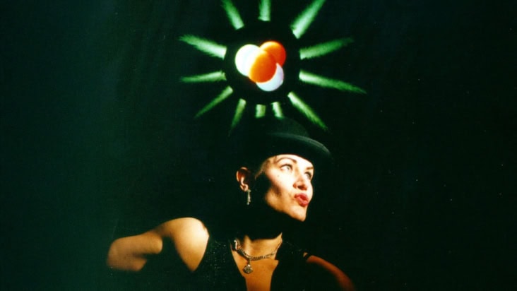Lynda Williams aka Physics Chanteuse on stage, wearing a black hat with a glowing atom graphic above her head