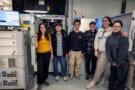 Six people stand side-by-side in a laboratory in front of a large device that resembles a 5-foot-tall refrigerator with microscope equipment inside the top drawer
