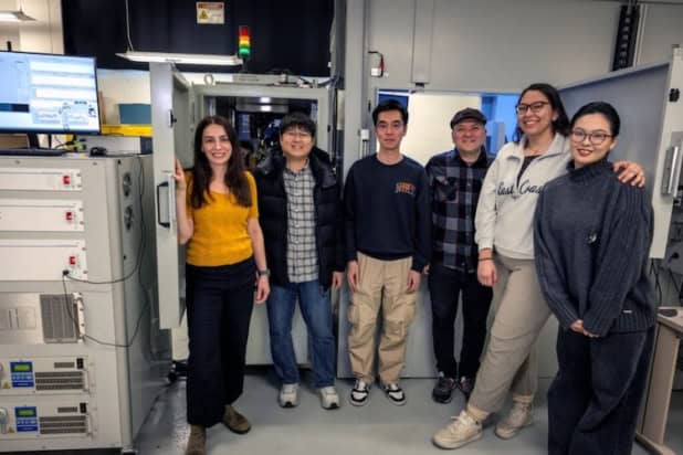 Six people stand side-by-side in a laboratory in front of a large device that resembles a 5-foot-tall refrigerator with microscope equipment inside the top drawer