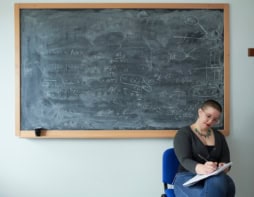 A woman sat writing in a notebook in front of a blackboard filled with equations