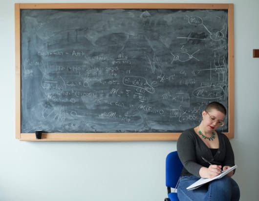 A woman sat writing in a notebook in front of a blackboard filled with equations