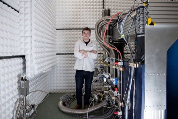 A photo of Joseph Carroll standing in a laboratory next to a large machine with many cables, pipes and valves running into it. He's wearing a white shirt and black jeans, and the walls of the lab are covered in white acoustic insulation material