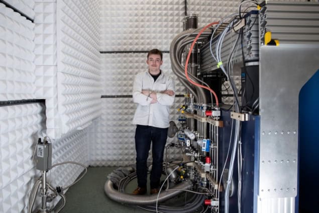 03-07-2025 joseph carroll superconducting stm web A photo of Joseph Carroll standing in a laboratory next to a large machine with many cables, pipes and valves running into it. He's wearing a white shirt and black jeans, and the walls of the lab are covered in white acoustic insulation material