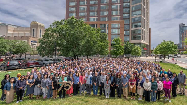 Large group of people stood on grass outside a tall building holding a "75" sign