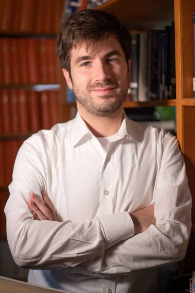 Photo of Christopher Tunnell standing in an office environment. He's wearing a white button-down shirt and there are bookcases in the background